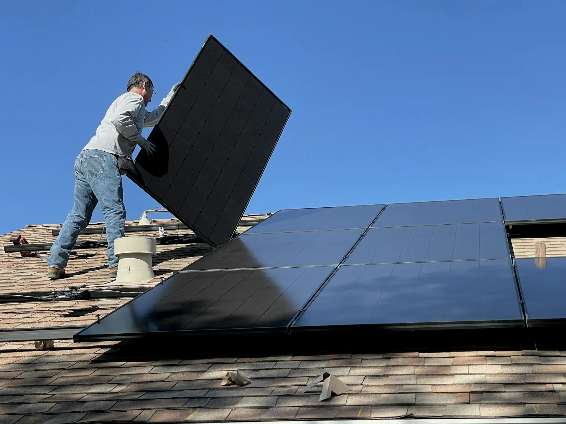 Industry context: A person installing a black solar panel on a shingled roof under blue sky